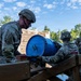A group of military personnel in camouflage uniforms and helmets are working together to maneuver a blue barrel marked with the number '4' across a wooden structure, likely as part of a team-building or obstacle course exercise. The setting appears to be an outdoor military training area with wooden barriers and lush green trees in the background. The activity demonstrates coordinated teamwork under a training scenario.