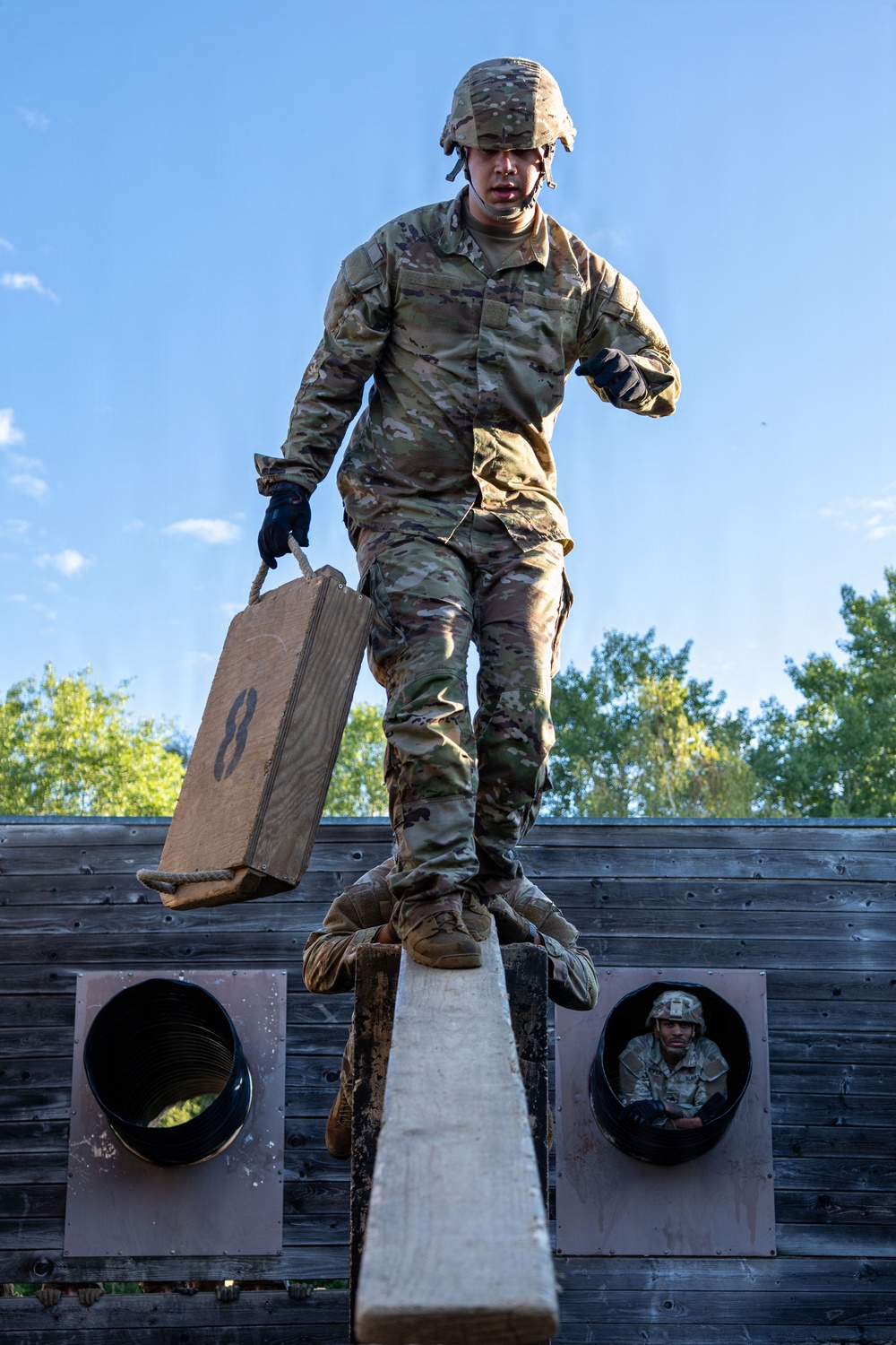 A soldier in full OCP (Operational Camouflage Pattern) uniform and combat helmet navigates a military obstacle course, balancing on a narrow beam while carrying a wooden ammunition crate marked with the number '8'. A second soldier is visible crawling through a circular tunnel opening in the wooden wall below. The image appears to be taken during a military training exercise under clear blue skies.