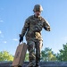 A soldier in full OCP (Operational Camouflage Pattern) uniform and combat helmet navigates a military obstacle course, balancing on a narrow beam while carrying a wooden ammunition crate marked with the number '8'. A second soldier is visible crawling through a circular tunnel opening in the wooden wall below. The image appears to be taken during a military training exercise under clear blue skies.