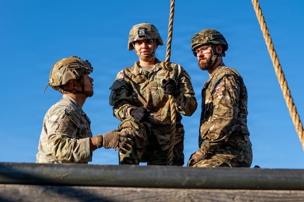 Three military personnel in camouflage uniforms and helmets are positioned at what appears to be an elevated obstacle course platform. The central figure is holding a thick rope, suggesting a rope-climbing or rappelling exercise. The soldiers appear to be engaged in training activity, with two observing and one actively handling the rope.