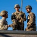 Three military personnel in camouflage uniforms and helmets are positioned at what appears to be an elevated obstacle course platform. The central figure is holding a thick rope, suggesting a rope-climbing or rappelling exercise. The soldiers appear to be engaged in training activity, with two observing and one actively handling the rope.