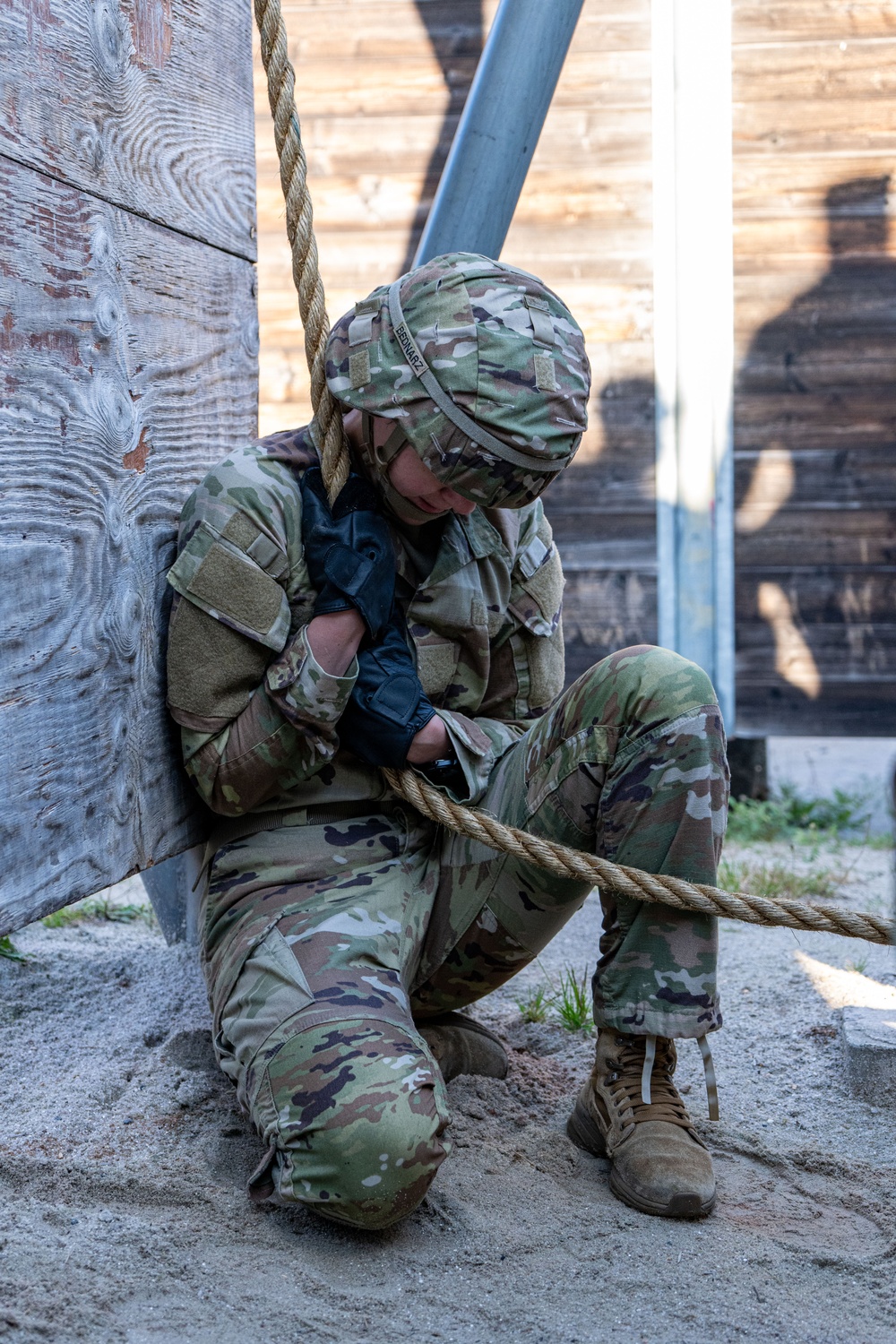 A soldier in full MultiCam/OCP camouflage uniform and helmet is seated on sandy ground against a weathered wooden wall, with a thick rope draped over their shoulder and across their body. The individual appears to be resting or recovering, with head bowed downward. A name tape reading 'BEDNARZ' is visible on the helmet cover.
