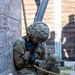 A soldier in full MultiCam/OCP camouflage uniform and helmet is seated on sandy ground against a weathered wooden wall, with a thick rope draped over their shoulder and across their body. The individual appears to be resting or recovering, with head bowed downward. A name tape reading 'BEDNARZ' is visible on the helmet cover.