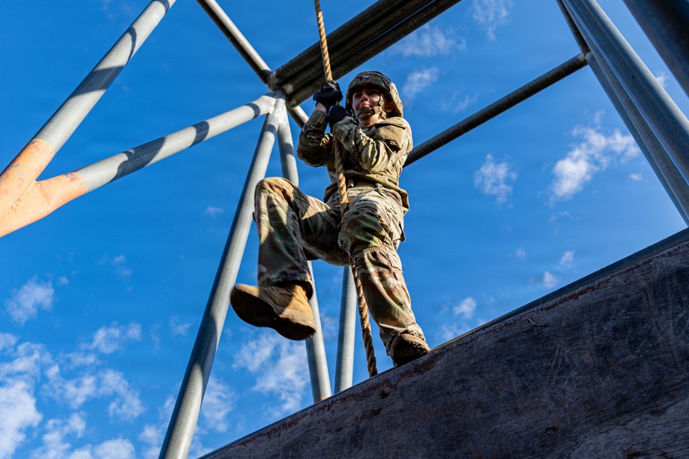 A soldier in full camouflage uniform and helmet is climbing or descending a thick rope on a military obstacle course or training tower. The photo is taken from a low angle looking upward, emphasizing the height and physical challenge of the activity. The structure consists of metal scaffolding with a wooden wall element, set against a bright blue sky with scattered clouds.