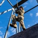 A soldier in full camouflage uniform and helmet is climbing or descending a thick rope on a military obstacle course or training tower. The photo is taken from a low angle looking upward, emphasizing the height and physical challenge of the activity. The structure consists of metal scaffolding with a wooden wall element, set against a bright blue sky with scattered clouds.