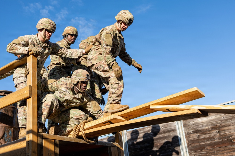 Four soldiers in US Army OCP (Operational Camouflage Pattern) uniforms are working together on a wooden obstacle course structure. They appear to be engaged in a team-building or combat training exercise, with one soldier crouching while holding a rope and others assisting or navigating the wooden framework. The image is taken from a low angle against a clear blue sky.
