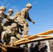 Four soldiers in US Army OCP (Operational Camouflage Pattern) uniforms are working together on a wooden obstacle course structure. They appear to be engaged in a team-building or combat training exercise, with one soldier crouching while holding a rope and others assisting or navigating the wooden framework. The image is taken from a low angle against a clear blue sky.