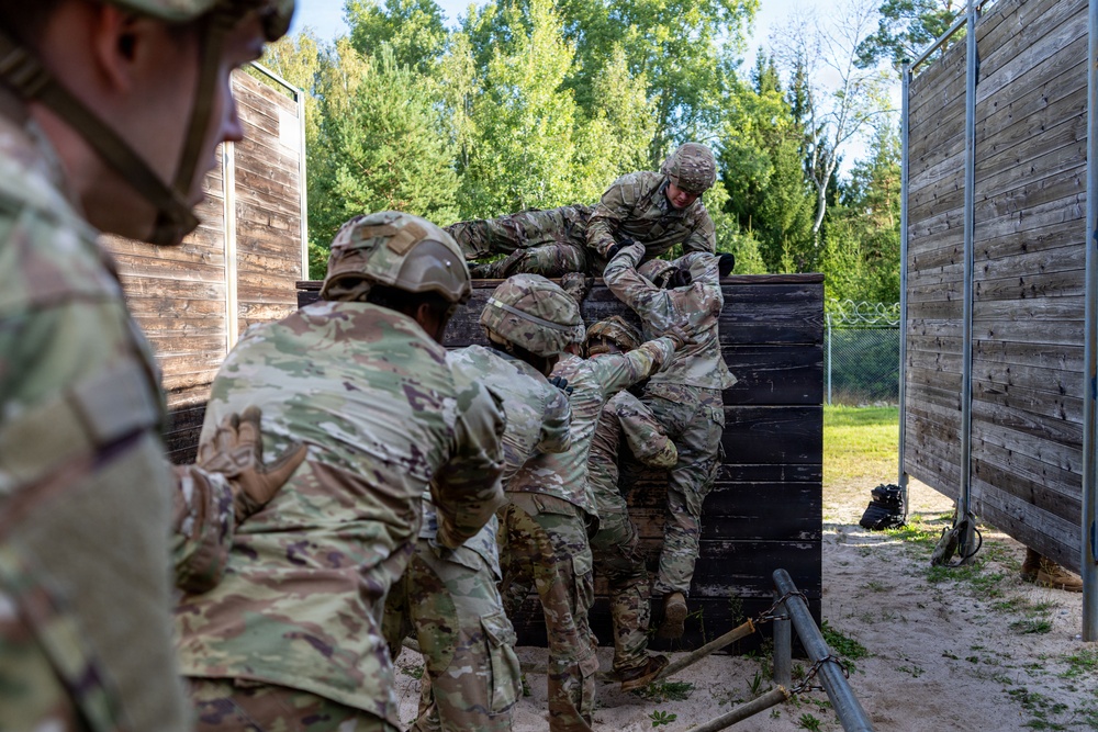 A group of military personnel in camouflage uniforms and helmets are working together to scale or assist others over a tall wooden wall obstacle. The scene appears to take place on a military training course, with sandy ground, chain-link fencing, and wooden barrier structures visible. The teamwork-focused activity suggests a unit cohesion or obstacle course training exercise.