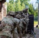 A group of military personnel in camouflage uniforms and helmets are working together to scale or assist others over a tall wooden wall obstacle. The scene appears to take place on a military training course, with sandy ground, chain-link fencing, and wooden barrier structures visible. The teamwork-focused activity suggests a unit cohesion or obstacle course training exercise.