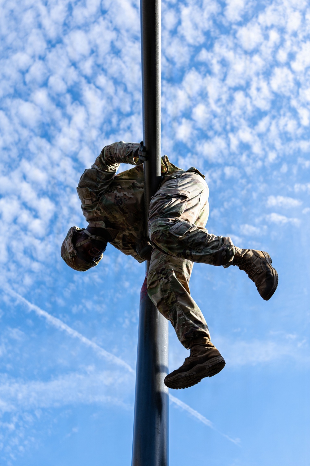 A military service member in full camouflage uniform is climbing a vertical metal pole, photographed from a low angle looking upward against a partly cloudy blue sky. The soldier is mid-climb, with arms gripping the pole and legs wrapped around it in a classic pole-climbing technique. The dramatic upward perspective emphasizes the height and physical effort involved in the activity.