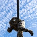 A military service member in full camouflage uniform is climbing a vertical metal pole, photographed from a low angle looking upward against a partly cloudy blue sky. The soldier is mid-climb, with arms gripping the pole and legs wrapped around it in a classic pole-climbing technique. The dramatic upward perspective emphasizes the height and physical effort involved in the activity.