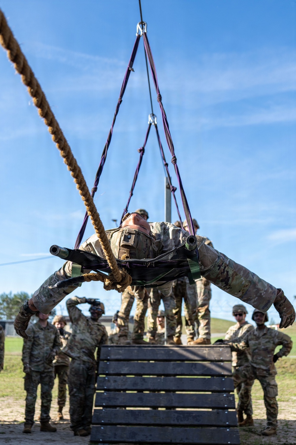 A military service member in full camouflage uniform and combat gear is suspended horizontally in a harness rigging system attached to an overhead structure, appearing to practice rappelling or sling-load procedures. Multiple other uniformed personnel in OCP (Operational Camouflage Pattern) uniforms observe from the ground level. The setting appears to be a military training facility with sandy ground and wooden platform structures.