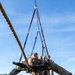 A military service member in full camouflage uniform and combat gear is suspended horizontally in a harness rigging system attached to an overhead structure, appearing to practice rappelling or sling-load procedures. Multiple other uniformed personnel in OCP (Operational Camouflage Pattern) uniforms observe from the ground level. The setting appears to be a military training facility with sandy ground and wooden platform structures.
