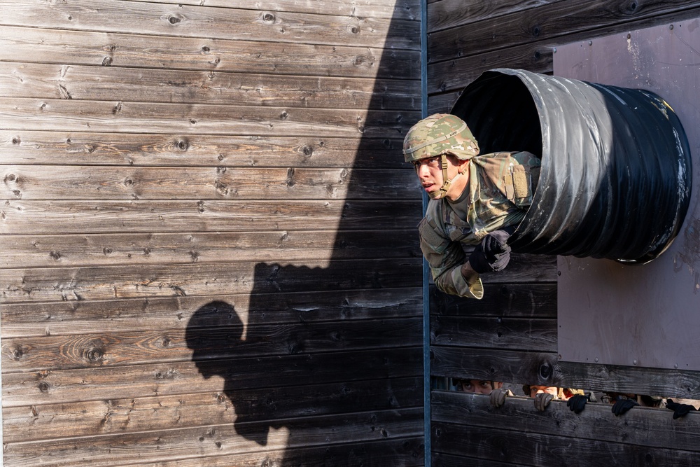 A soldier in full military camouflage uniform and helmet is emerging headfirst from a large black cylindrical tunnel mounted on a wooden wall structure, part of a military obstacle course. The image is taken from an elevated angle, capturing the soldier mid-crawl as they exit the tunnel. A shadow of an observer is cast on the weathered wooden wall to the left, and additional personnel are partially visible below.