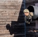 A soldier in full military camouflage uniform and helmet is emerging headfirst from a large black cylindrical tunnel mounted on a wooden wall structure, part of a military obstacle course. The image is taken from an elevated angle, capturing the soldier mid-crawl as they exit the tunnel. A shadow of an observer is cast on the weathered wooden wall to the left, and additional personnel are partially visible below.