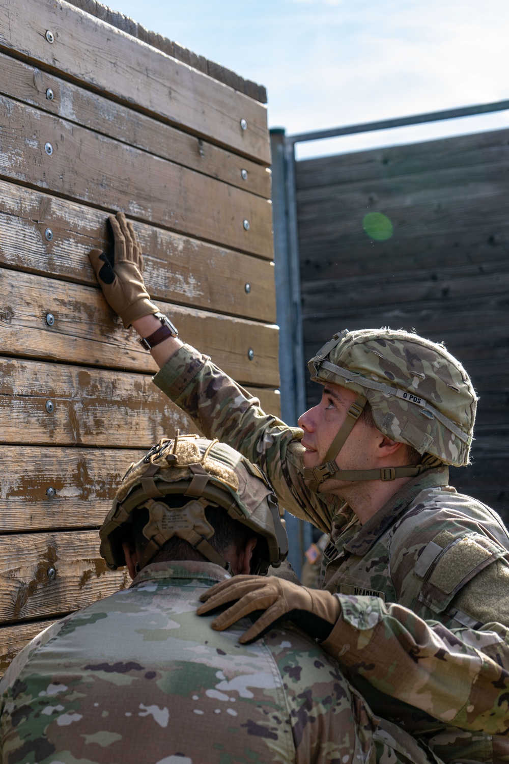 Two U.S. Army soldiers in full combat gear are working together to scale or navigate a tall wooden wall obstacle, consistent with a military training course. The soldier facing the camera is reaching upward with a gloved hand while using the other soldier as support. Both wear OCP (Operational Camouflage Pattern) uniforms and tactical helmets.