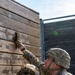 Two U.S. Army soldiers in full combat gear are working together to scale or navigate a tall wooden wall obstacle, consistent with a military training course. The soldier facing the camera is reaching upward with a gloved hand while using the other soldier as support. Both wear OCP (Operational Camouflage Pattern) uniforms and tactical helmets.