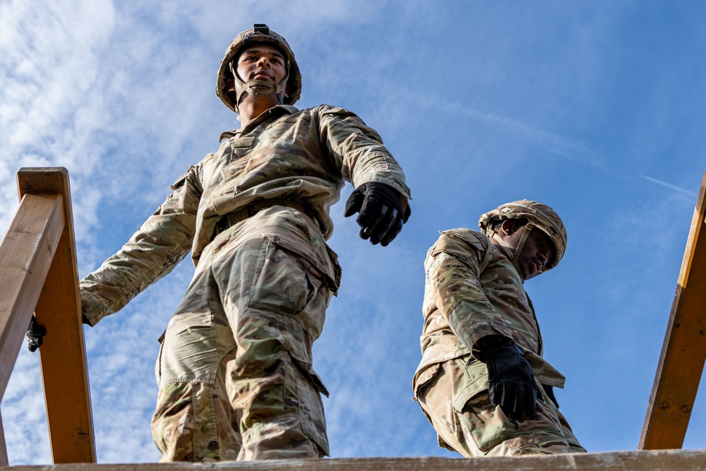 Two military personnel in full combat uniforms are photographed from a low-angle perspective, appearing to stand atop a wooden structure. Both individuals are wearing OCP (Operational Camouflage Pattern) uniforms, tactical helmets, and black gloves. The dramatic upward camera angle against a partly cloudy blue sky emphasizes their elevated position.