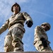 Two military personnel in full combat uniforms are photographed from a low-angle perspective, appearing to stand atop a wooden structure. Both individuals are wearing OCP (Operational Camouflage Pattern) uniforms, tactical helmets, and black gloves. The dramatic upward camera angle against a partly cloudy blue sky emphasizes their elevated position.