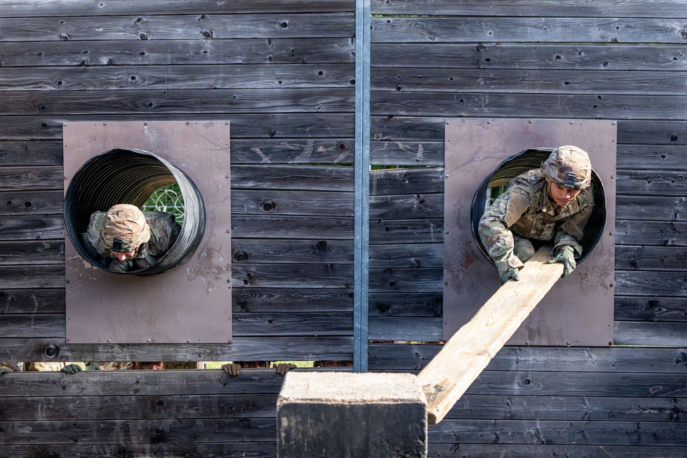 Two military personnel in camouflage uniforms and helmets are navigating a training obstacle course, crawling through circular tunnel openings in a dark wooden wall. One soldier on the right is actively pushing or maneuvering a wooden beam/plank through the opening. The image is taken from an elevated angle, providing a top-down perspective of the exercise.