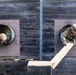Two military personnel in camouflage uniforms and helmets are navigating a training obstacle course, crawling through circular tunnel openings in a dark wooden wall. One soldier on the right is actively pushing or maneuvering a wooden beam/plank through the opening. The image is taken from an elevated angle, providing a top-down perspective of the exercise.