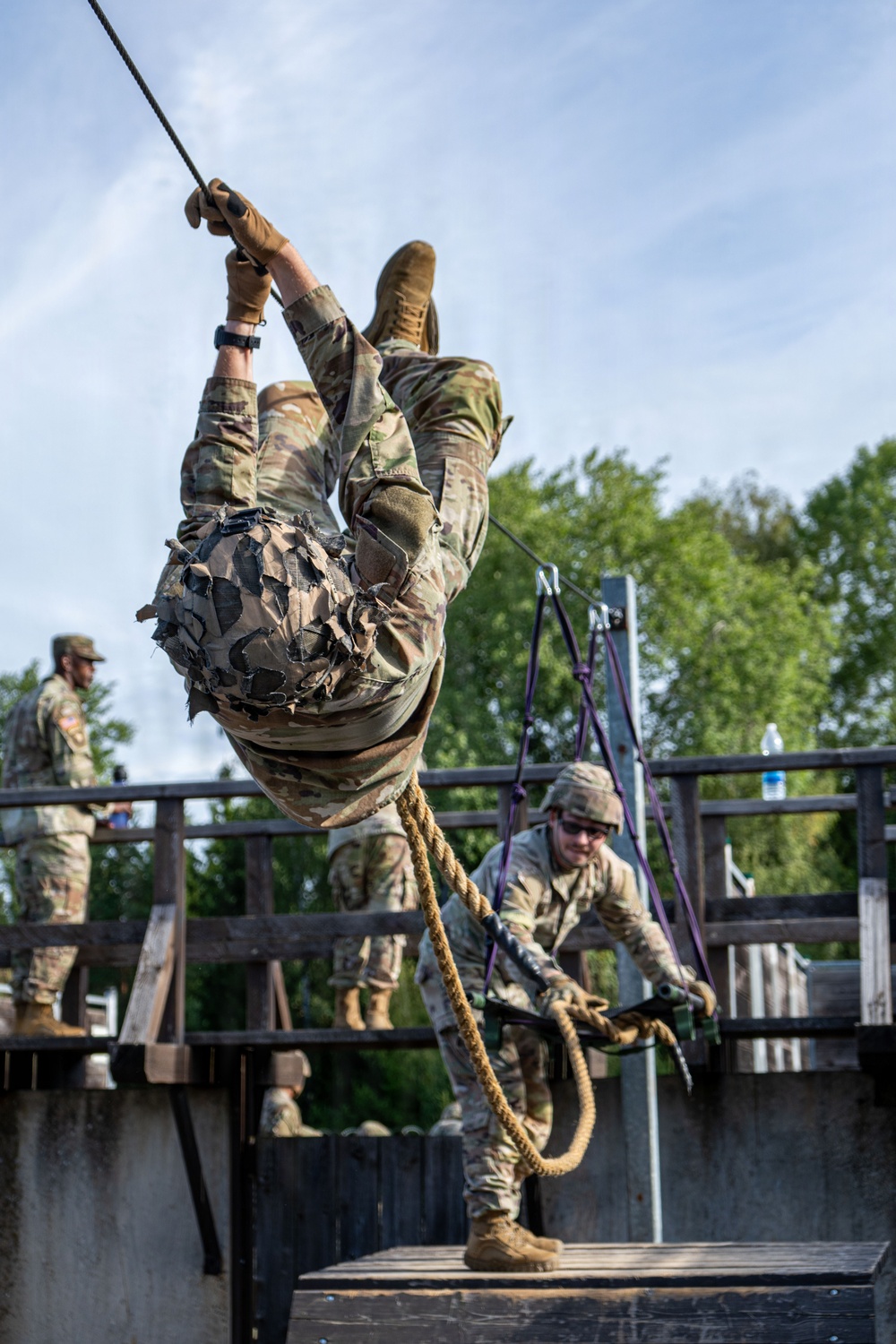 Military personnel are conducting a rope training exercise on a wooden platform structure. One soldier is suspended upside-down from a rope, wearing full camouflage uniform including a ghillie-style vest, while a second soldier on the platform manages the belay system. Additional uniformed personnel are visible observing in the background.