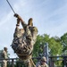 Military personnel are conducting a rope training exercise on a wooden platform structure. One soldier is suspended upside-down from a rope, wearing full camouflage uniform including a ghillie-style vest, while a second soldier on the platform manages the belay system. Additional uniformed personnel are visible observing in the background.
