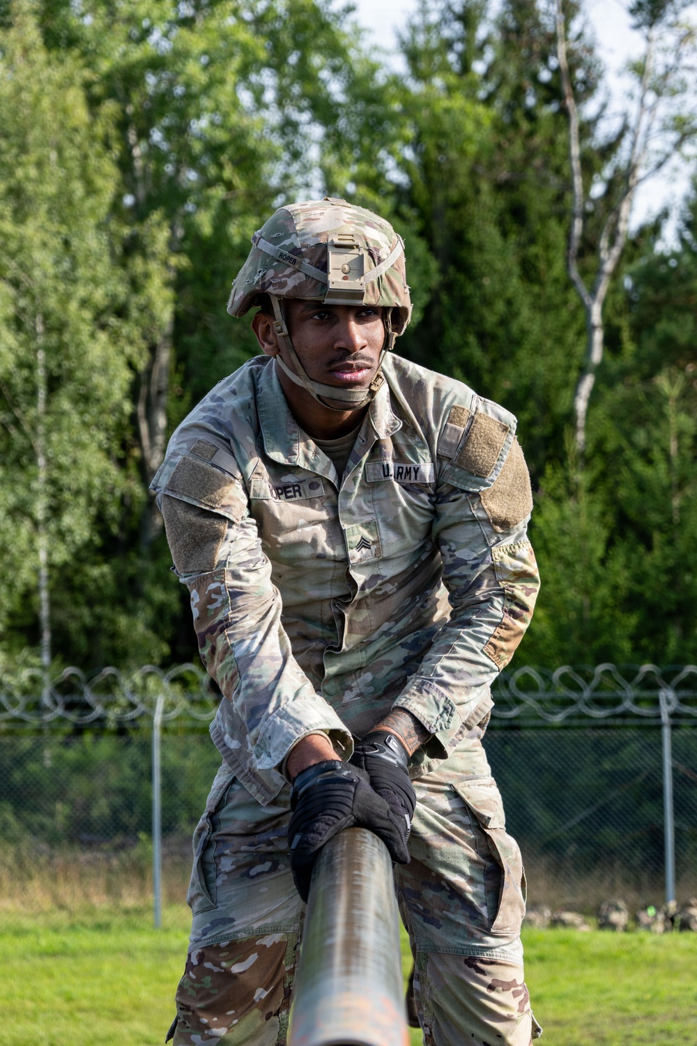 A U.S. Army soldier in full OCP (Operational Camouflage Pattern) uniform and combat helmet is navigating what appears to be an obstacle course, leaning over a horizontal bar or beam. The soldier wears tactical gloves and has a name tape partially visible reading '...PER' and a 'U.S. ARMY' chest tape. The background shows a green, wooded outdoor training area with a chain-link fence topped with concertina wire.