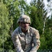 A U.S. Army soldier in full OCP (Operational Camouflage Pattern) uniform and combat helmet is navigating what appears to be an obstacle course, leaning over a horizontal bar or beam. The soldier wears tactical gloves and has a name tape partially visible reading '...PER' and a 'U.S. ARMY' chest tape. The background shows a green, wooded outdoor training area with a chain-link fence topped with concertina wire.