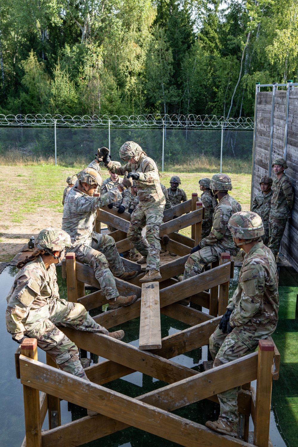 A group of military personnel in camouflage uniforms and helmets are navigating a wooden obstacle course structure over what appears to be a water pit. The soldiers are working cooperatively to traverse a multi-level wooden lattice/grid obstacle, with some assisting others across the beams. The setting appears to be a military training facility with a chain-link fence topped with concertina wire visible in the background.