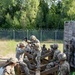 A group of military personnel in camouflage uniforms and helmets are navigating a wooden obstacle course structure over what appears to be a water pit. The soldiers are working cooperatively to traverse a multi-level wooden lattice/grid obstacle, with some assisting others across the beams. The setting appears to be a military training facility with a chain-link fence topped with concertina wire visible in the background.