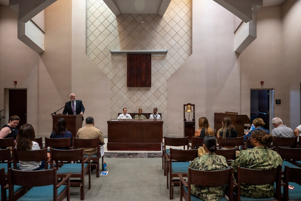 A formal ceremony or meeting is taking place in what appears to be a military chapel or multi-purpose ceremonial space. A civilian man in a suit is speaking at a podium on the left, while three uniformed military personnel are seated at a table at the front. Attendees, including both civilians and military personnel in Navy uniforms, are seated in wooden chairs with teal cushions.