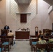 A formal ceremony or meeting is taking place in what appears to be a military chapel or multi-purpose ceremonial space. A civilian man in a suit is speaking at a podium on the left, while three uniformed military personnel are seated at a table at the front. Attendees, including both civilians and military personnel in Navy uniforms, are seated in wooden chairs with teal cushions.
