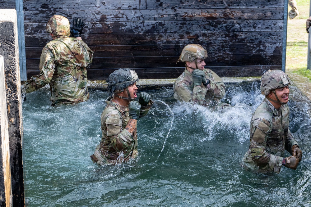 Four military personnel in full combat uniforms and helmets are navigating a water obstacle during what appears to be a military training exercise or obstacle course. The soldiers are wading through a water pit, with one visibly shouting or reacting to the cold water. The wooden wall structure behind them suggests this is a purpose-built military training facility.