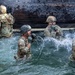 Four military personnel in full combat uniforms and helmets are navigating a water obstacle during what appears to be a military training exercise or obstacle course. The soldiers are wading through a water pit, with one visibly shouting or reacting to the cold water. The wooden wall structure behind them suggests this is a purpose-built military training facility.