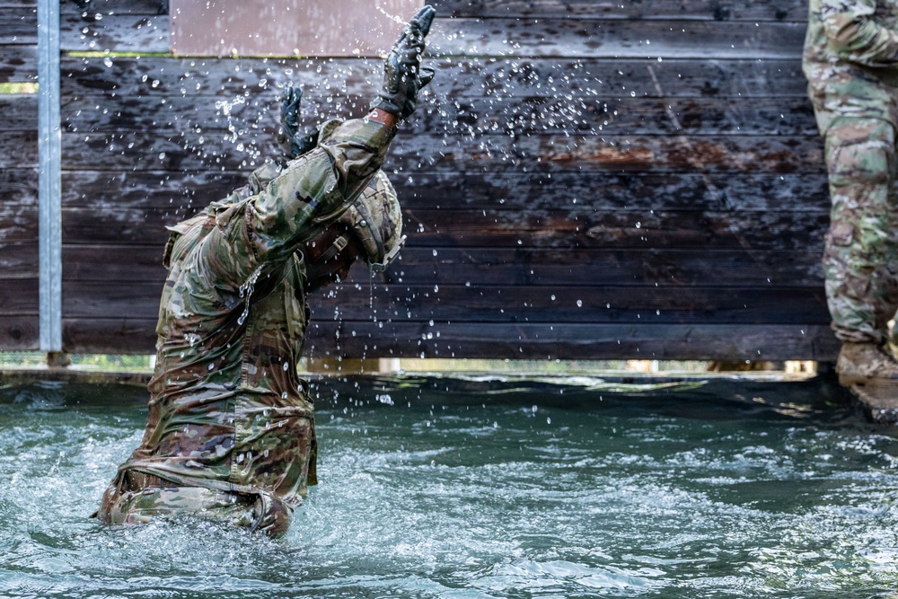 A soldier in full camouflage uniform and tactical gloves is emerging from or moving through a water obstacle, with water splashing dramatically around them. The individual appears to be participating in a military training exercise involving a water crossing or obstacle course. A second soldier in matching camouflage is partially visible on the right edge of the frame.