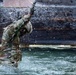A soldier in full camouflage uniform and tactical gloves is emerging from or moving through a water obstacle, with water splashing dramatically around them. The individual appears to be participating in a military training exercise involving a water crossing or obstacle course. A second soldier in matching camouflage is partially visible on the right edge of the frame.