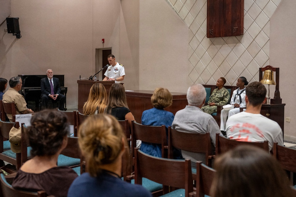A military ceremony or formal event is taking place in what appears to be a chapel or auditorium on a military installation. A naval officer in white dress uniform is speaking at a podium, while an audience of civilians and military personnel are seated in pews. Several uniformed military members are seated on the stage area alongside a civilian dignitary.