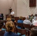A military ceremony or formal event is taking place in what appears to be a chapel or auditorium on a military installation. A naval officer in white dress uniform is speaking at a podium, while an audience of civilians and military personnel are seated in pews. Several uniformed military members are seated on the stage area alongside a civilian dignitary.