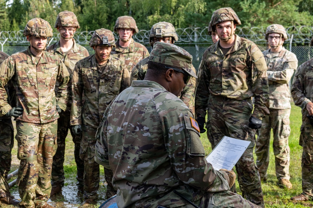 A military instructor or non-commissioned officer, wearing a patrol cap and US Army OCP (Operational Camouflage Pattern) uniform with a US flag patch, is kneeling and reading from a document while addressing a group of soldiers. The soldiers are standing in a semicircle, wearing helmets and OCP uniforms, attentively listening during what appears to be a field briefing or training exercise. The setting is an outdoor military training area with a chain-link fence topped with concertina wire visible in the background.