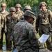 A military instructor or non-commissioned officer, wearing a patrol cap and US Army OCP (Operational Camouflage Pattern) uniform with a US flag patch, is kneeling and reading from a document while addressing a group of soldiers. The soldiers are standing in a semicircle, wearing helmets and OCP uniforms, attentively listening during what appears to be a field briefing or training exercise. The setting is an outdoor military training area with a chain-link fence topped with concertina wire visible in the background.