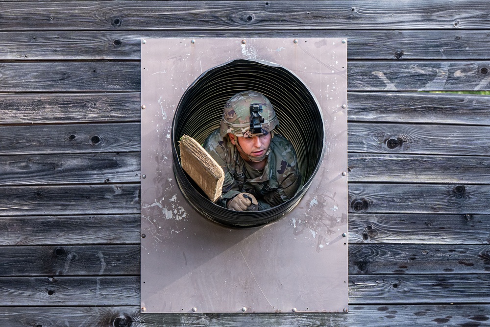 A soldier in military camouflage uniform and helmet is crawling through a circular tunnel obstacle embedded in a wooden wall. The individual appears to be navigating a military obstacle course or training exercise. The photo is taken from a straight-on perspective, framing the soldier within the circular tunnel opening.