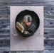 A soldier in military camouflage uniform and helmet is crawling through a circular tunnel obstacle embedded in a wooden wall. The individual appears to be navigating a military obstacle course or training exercise. The photo is taken from a straight-on perspective, framing the soldier within the circular tunnel opening.
