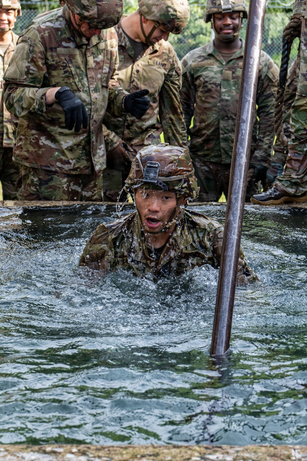 A U.S. Army soldier in full combat uniform and helmet is emerging from a water obstacle during what appears to be a military training exercise or obstacle course. Several other soldiers in OCP (Operational Camouflage Pattern) uniforms and helmets observe and provide guidance from the edge of the water pit. The soldier in the water is visibly soaked and appears to be exerting significant effort.
