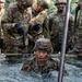 A U.S. Army soldier in full combat uniform and helmet is emerging from a water obstacle during what appears to be a military training exercise or obstacle course. Several other soldiers in OCP (Operational Camouflage Pattern) uniforms and helmets observe and provide guidance from the edge of the water pit. The soldier in the water is visibly soaked and appears to be exerting significant effort.