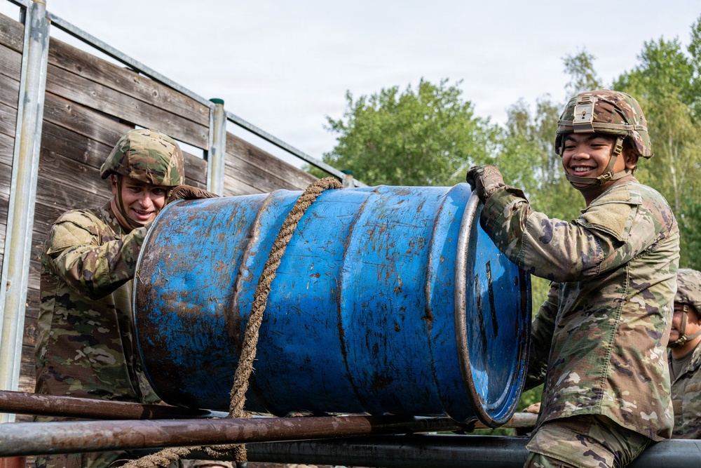 Two soldiers in camouflage uniforms and helmets are working together to lift or maneuver a large, mud-covered blue metal barrel on what appears to be the bed of a military truck or flatbed vehicle. A third soldier is partially visible in the background. The scene appears to be a training exercise or field operation, with one soldier visibly smiling.