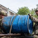Two soldiers in camouflage uniforms and helmets are working together to lift or maneuver a large, mud-covered blue metal barrel on what appears to be the bed of a military truck or flatbed vehicle. A third soldier is partially visible in the background. The scene appears to be a training exercise or field operation, with one soldier visibly smiling.