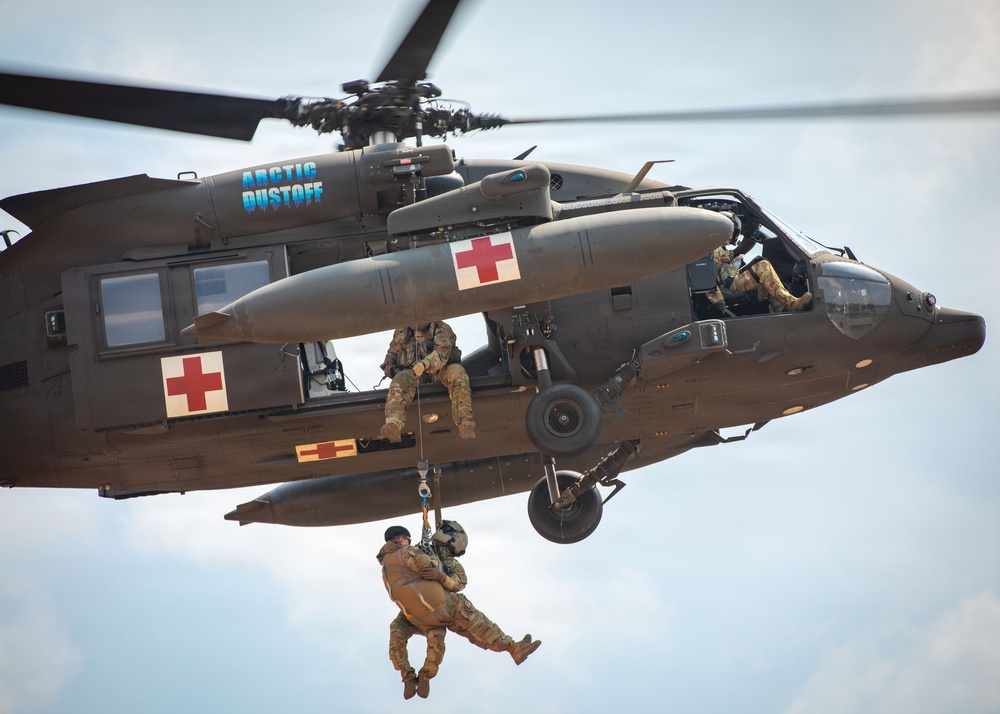 A military medical evacuation (MEDEVAC) helicopter marked 'Arctic Dustoff' performs a hoist rescue operation in flight. Two personnel are suspended below the aircraft on a hoist cable, with one appearing to hold or rescue the other. A crew member is visible in the open side door managing the operation while pilots are visible in the cockpit.