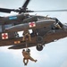 A military medical evacuation (MEDEVAC) helicopter marked 'Arctic Dustoff' performs a hoist rescue operation in flight. Two personnel are suspended below the aircraft on a hoist cable, with one appearing to hold or rescue the other. A crew member is visible in the open side door managing the operation while pilots are visible in the cockpit.