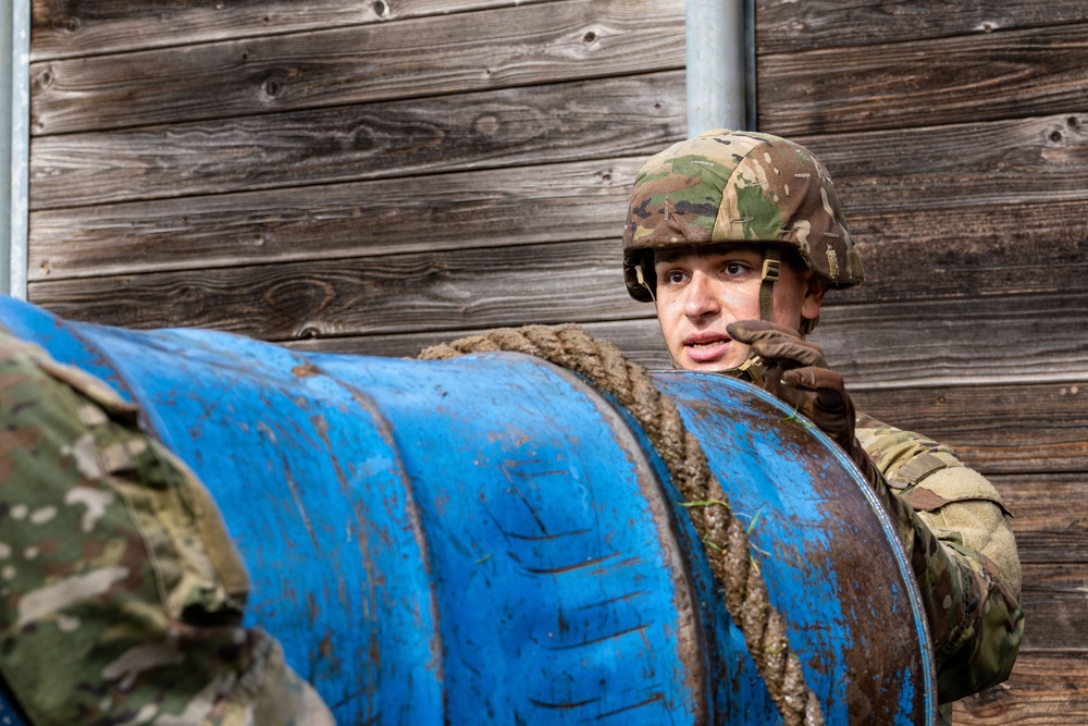 A soldier in camouflage uniform and helmet is handling a large blue barrel or drum covered in mud and rope. The soldier appears to be participating in a military training exercise, likely an obstacle course or physical training drill. A wooden plank wall serves as the background.